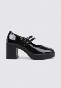 Zapatos de tacón de charol negro con un tacón grueso, que presentan dobles correas con hebillas circulares. Superficie lisa y diseño de punta redondeada.