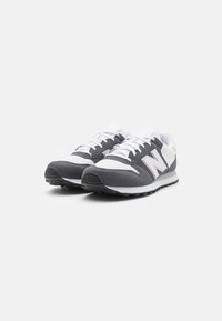 Szaro-białe trampki z siateczki i zamszu, z zaokrąglonym noskiem i fakturowaną podeszwą. Po boku znajduje się wyraźne logo.