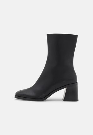 Bottine noire avec une finition en cuir synthétique lisse, bout carré, et talon bloc noir nervuré avec un accent argenté.