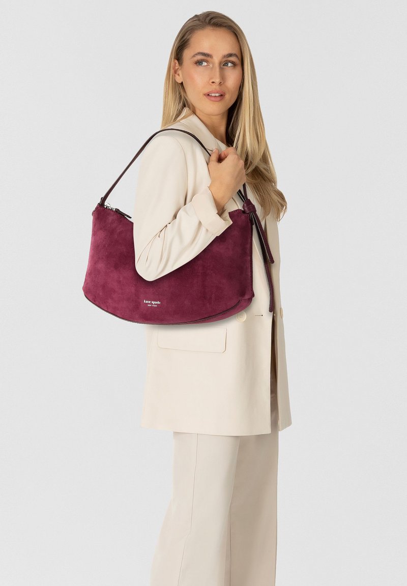 Borsa a tracolla in suede bordeaux con forma curva, tracolla in pelle e dettaglio logo sul fondo. Abbinata a un outfit su misura color crema.