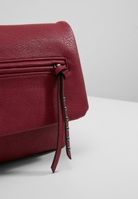 Červená kožená taška s texturovaným povrchem, vybavená kapsou na zip a černým popruhem zdobeným kovovými nýty.