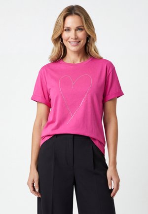 Uśmiechnięta kobieta o blond włosach do ramion, ubrana w różowy t-shirt z wzorem w kropki w kształcie serca oraz czarne spodnie na białym tle.