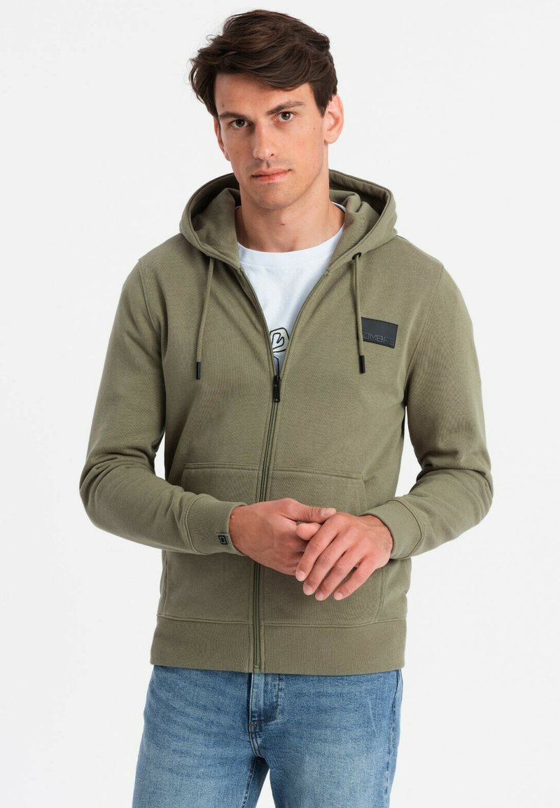 Sweat à capuche zippé vert olive avec une texture lisse, capuche à cordon, poches avant et un petit patch logo noir sur la poitrine. Un jean est porté en dessous.