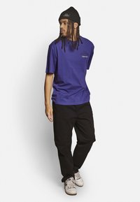 Lila T-Shirt mit kurzen Ärmeln und Textlogo, kombiniert mit schwarzen Hosen und einer schwarzen Mütze. Das Modell steht in einer neutralen Pose mit Sneakers.