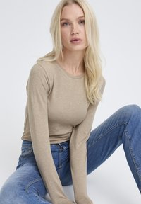 Blonde Frau mit loose Haaren, die ein beiges Langarmoberteil und blaue Jeans trägt, sitzt mit einem Knie hoch gegen einen weißen Hintergrund.