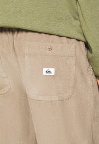 Pantalon en velours côtelé beige avec poche arrière boutonnée ornée d'un écusson logo Quiksilver, associé à un haut vert olive.