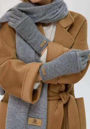 Guanti in maglia grigia con un'etichetta logo, indossati con una sciarpa grigia chiara e un cappotto in pile beige. I guanti hanno una vestibilità aderente e un tessuto testurizzato.