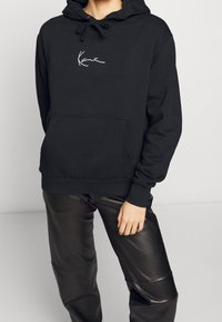 Czarna bluza z kapturem z mieszanki bawełny z kieszenią kangurką i białym haftowanym logo, w połączeniu z czarnymi skórzanymi spodniami.