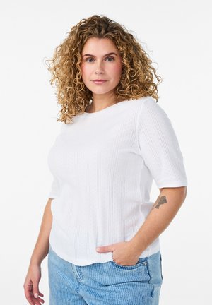 Kvinde med krøllet blondt hår iført en hvid tekstureret kortærmet top og lyseblå jeans, med en tatovering på venstre underarm.
