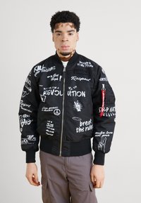 Black bomber jacket with white printed text and graphics, featuring a zip front, ribbed cuffs, and a red tab. Worn with gray pants.