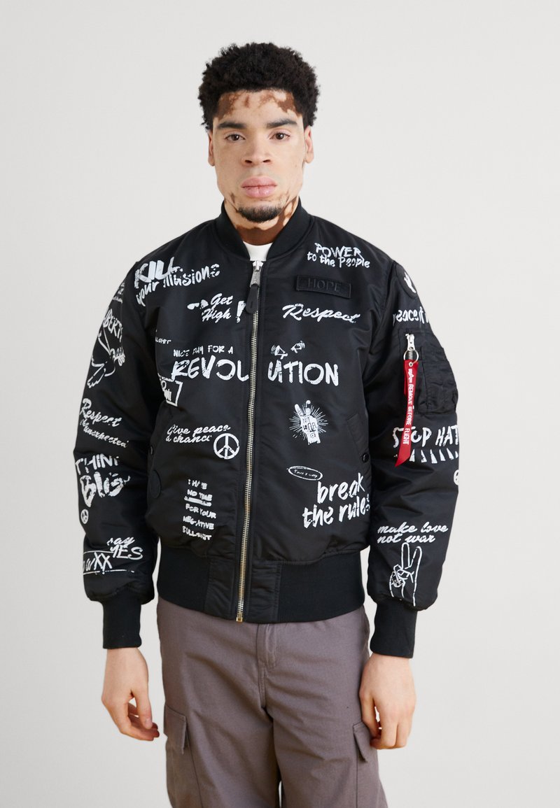 Black bomber jacket with white printed text and graphics, featuring a zip front, ribbed cuffs, and a red tab. Worn with gray pants.