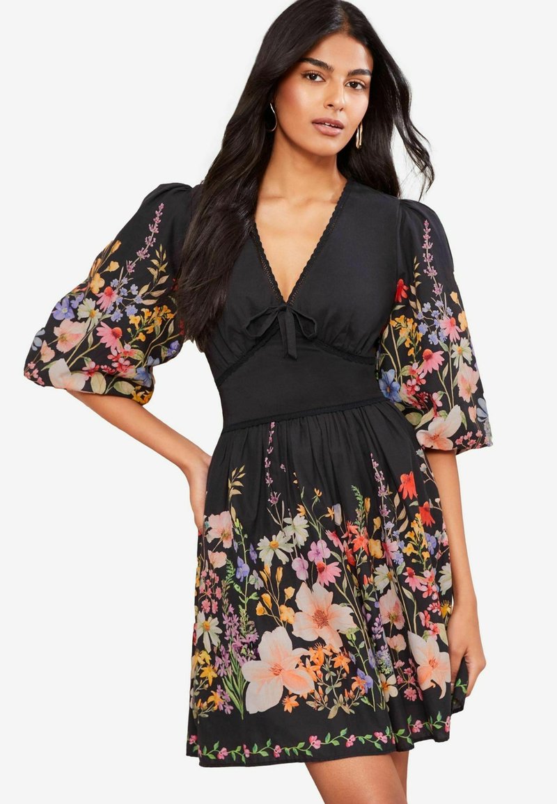 Robe florale noire avec manches bouffantes, présentant des imprimés de fleurs colorés dans diverses teintes. Décolleté en V avec un détail noué à l'avant.