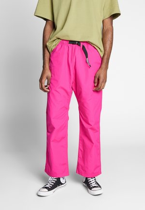 Mann trägt leuchtend pinke Hose mit schwarzem Gürtel, hellgrünes übergroßes T-Shirt und schwarze High-Top-Sneaker vor weißem Hintergrund.