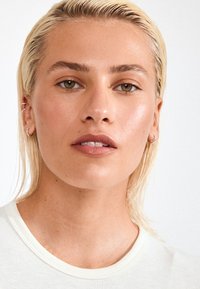 Světle barevné, jemně strukturované tričko s kulatým výstřihem. Osoba má krátké blond vlasy a na uších malé stříbrné kruhové náušnice. Hladký vzhled pleti.