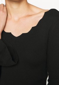 Haut noir en maille côtelée avec un col à bord festonné et des manches longues évasées, mettant en valeur un tissu texturé et une coupe ajustée.