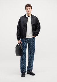 Chaqueta bomber negra, camiseta polo crema, pantalones azules con estampado geométrico, calzado negro y un bolso negro pequeño. Los pantalones presentan un diseño geométrico.