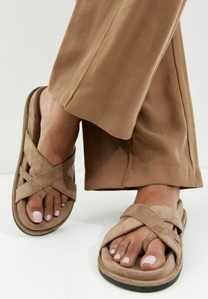 Pies con sandalias beige de correas cruzadas combinadas con pantalones marrones de pierna ancha, mostrando uñas de los pies pintadas de rosa claro.