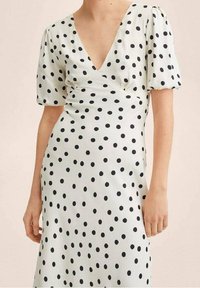 Robe à pois en tissu blanc avec des pois noirs, un décolleté en V profond et des manches courtes bouffantes, créant une silhouette ajustée.