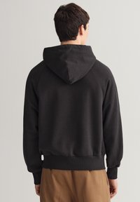 Černá mikina s kapucí na stažení, žebrovanými manžetami a lemem, vyrobená z měkkého materiálu. Má jednoduchý, bezspojový design bez vzorů.