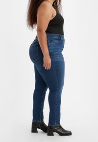 Úzké džíny v tmavě modrém denimu, vysoce pasované s minimálním poškozením, doplněné černými kotníkovými botami s hladkým povrchem.