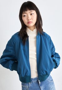 Chaqueta bomber color azul teal con puños y dobladillo acanalados, cierre de cremallera y tela ligera. Llevada sobre una blusa color crema, con un diseño de corte relajado.