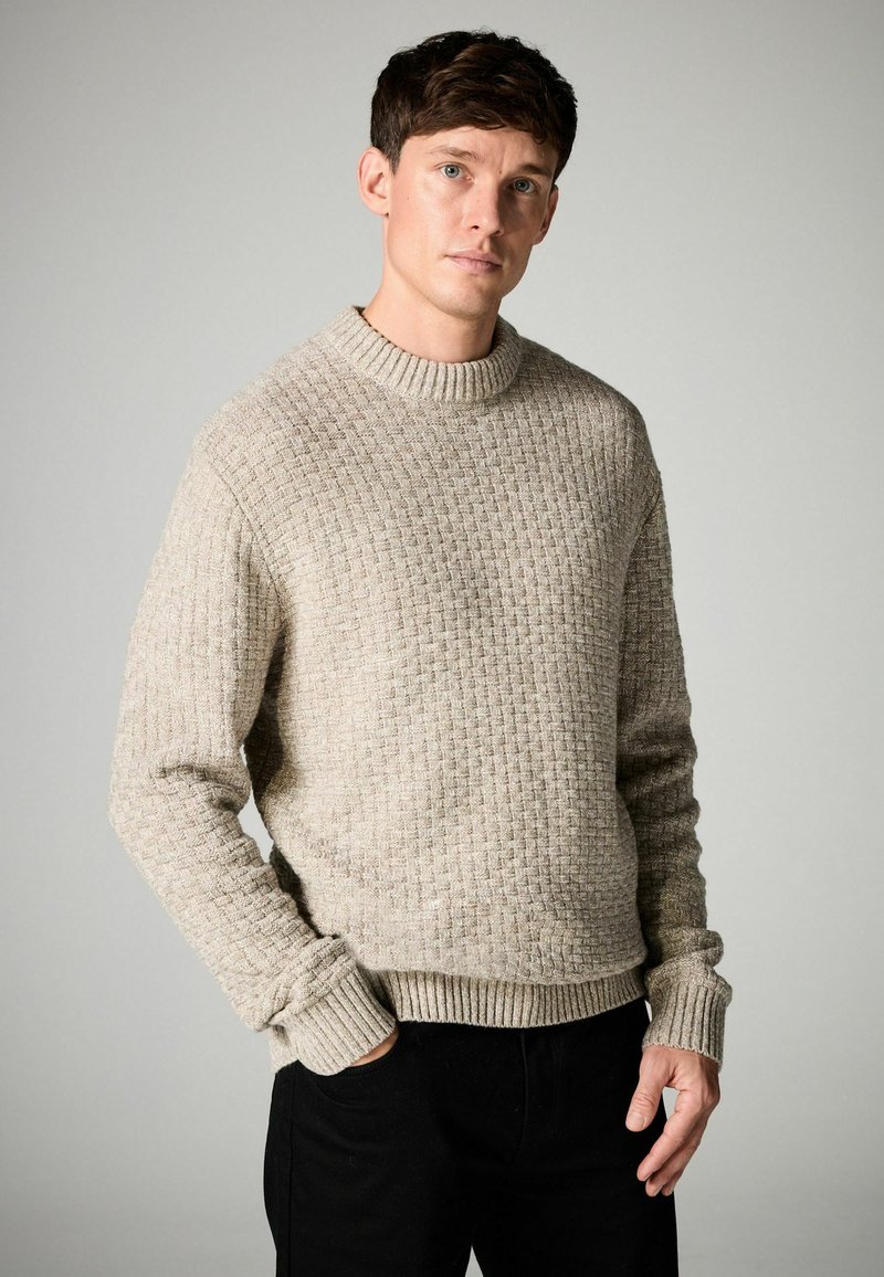 Beige knitted sweater with a textured pattern, ribbed crew neck, and cuffs, paired with black pants.