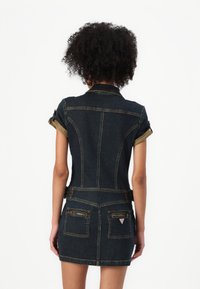 Kvinna med lockigt hår som bär en figursydd mörk jeansklänning med uppvikta ärmar och dragkedjefickor på baksidan, stående mot en neutral vit bakgrund.