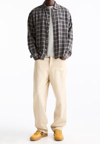 Personne portant un pantalon large beige, des baskets jaunes, un t-shirt gris et une chemise à carreaux marron boutonnée, les mains dans les poches.