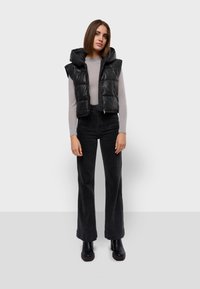 Gilet imbottito nero con cappuccio, caratterizzato da una chiusura con zip e senza maniche, indossato sopra un maglione aderente grigio chiaro e jeans neri a vita alta.