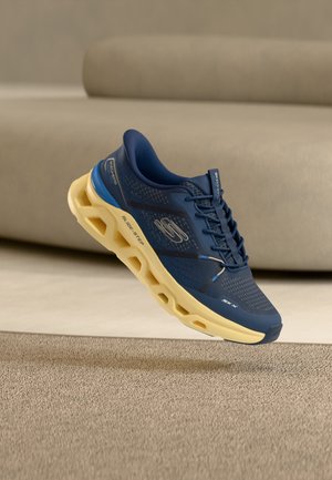 Chaussure de sport Skechers bleu marine avec semelle creuse hexagonale beige flottant au-dessus d'un tapis beige dans une pièce aux tons neutres.