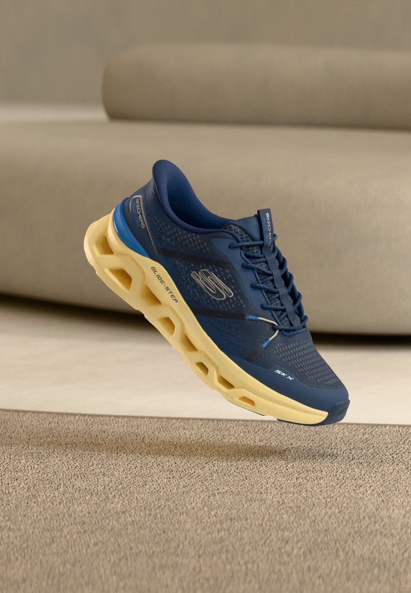 Ναυτικό μπλε αθλητικό παπούτσι Skechers με μπεζ, εξαγωνική κενή σόλα να αιωρείται πάνω από μπεζ χαλί σε δωμάτιο με ουδέτερους τόνους.