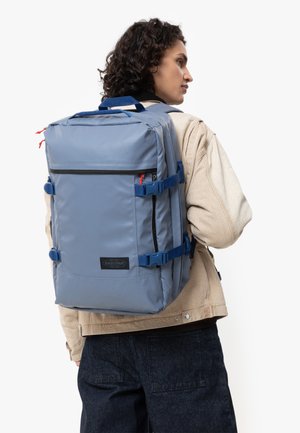 Sac à dos en tissu bleu avec plusieurs sangles et zippers, featuring un panneau avant et des poches latérales. Matériau durable avec une forme structurée.