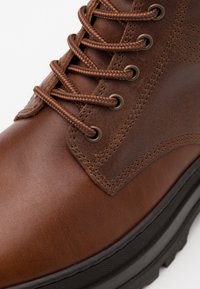 Bottine en cuir marron avec des lacets tressés marron, œillets en métal, texture lisse, semelle en caoutchouc noir contrastante et coutures renforcées.