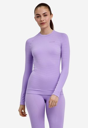 Kobieta stoi, ubrana w dopasowany top z długim rękawem i legginsy w jasnofioletowym stroju sportowym z logo na piersi na jednolitym tle.