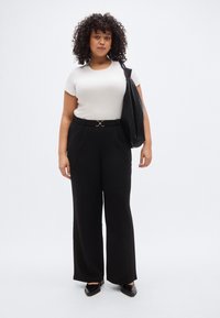 Μαύρο παντελόνι με φαρδιά μπατζάκια και χρυσή πόρπη ζώνης, συνδυασμένο με ένα εφαρμοστό κρεμ T-shirt. Μια μαύρη υφασμάτινη τσάντα ώμου κρέμεται από τον έναν ώμο.