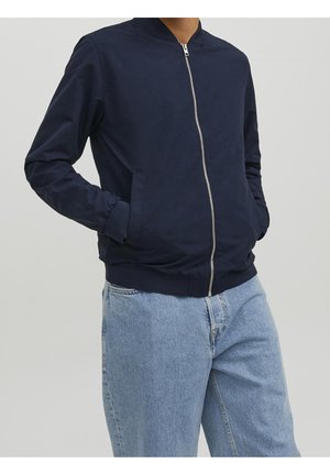 Mężczyzna w granatowej kurtce bomber na zamek z rękami w kieszeniach i jasnoniebieskich dżinsach na jednolitym tle.