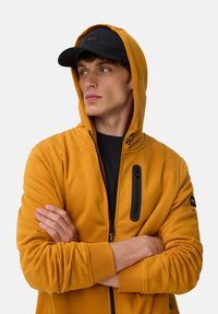 Jeune homme portant un sweat à capuche jaune moutarde avec une poche zippée noire et une casquette noire, les bras croisés, regardant sur le côté devant un fond uni.