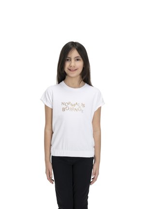 Une jeune fille souriante se tient devant un fond blanc. Elle porte un T-shirt blanc sur lequel est écrit « La normalité, c’est ennuyeux » et un pantalon noir.