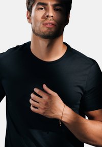 Svart t-shirt med kort ärm och rund halsringning. Modellens hand vilar på bröstet och visar upp ett tunt guldarmband med ett litet hänge.