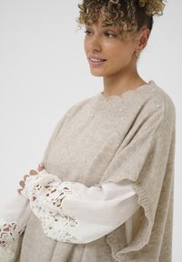 Pull en tricot beige avec des accents en perles, un col festonné, et des manches larges avec une finition en dentelle blanche et un tissu texturé.