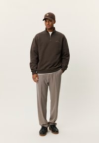 Καφέ φούτερ με φερμουάρ και ριμπ μανσέτες, συνδυασμένο με ριγέ παντελόνι σε μπεζ και καφέ. Μαύρα παπούτσια συμπληρώνουν την εμφάνιση.