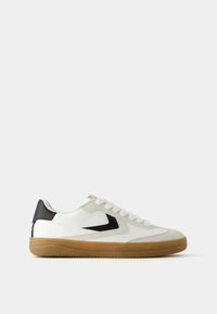 White low-top sneaker with beige suede accents, black heel patch, gum rubber sole, and white laces, shown in side profile on white background.