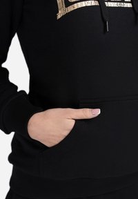 Black sweatshirt with a front pocket, textured fabric, and gold graphic detail. Features a drawstring hood and ribbed cuffs.