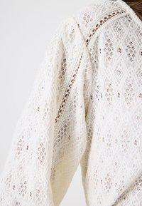 Tela de encaje blanca con intrincados patrones de diamantes y flores, mostrando una vista parcial de la manga y el hombro de la prenda sobre un fondo claro.