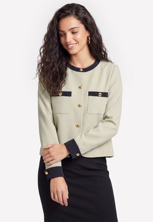 Kvinde med krøllet mørkt hår iført en beige jakke med sort kant og guldknapper, smilende og kigger ned med korslagte arme.