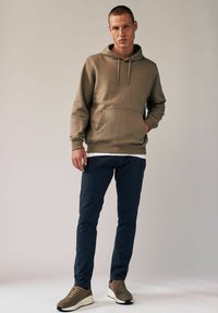 Beige Kapuzenpullover mit einer vorderen Tasche und Kordelzug, kombiniert mit navyblauen Slim-Fit-Hosen und braunen Sneakers mit hellen Akzenten. Glatte Stoffoberfläche.