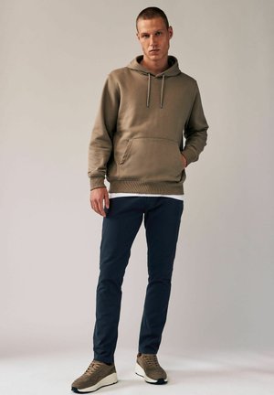 Felpa con cappuccio color sabbia dotata di tasca frontale e cordoncino, abbinata a pantaloni slim-fit blu scuro e sneakers marroni con dettagli chiari. Texture del tessuto liscia.