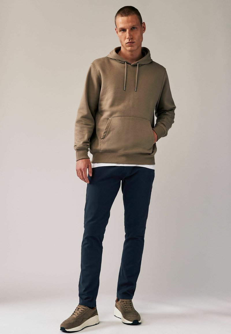 Beige Kapuzenpullover mit einer vorderen Tasche und Kordelzug, kombiniert mit navyblauen Slim-Fit-Hosen und braunen Sneakers mit hellen Akzenten. Glatte Stoffoberfläche.
