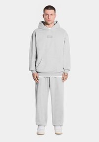 Młody mężczyzna stoi przodem, ubrany w jasnoszary bluzę z kapturem i dresy, trzymając ręce wzdłuż ciała.