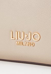 Beeži tekstuuriga nahkrahakott, millel on reljeefne kuldne "LIU·JO MILANO" logo. Sile viimistlus, struktureeritud kuju ja peened aktsendid.